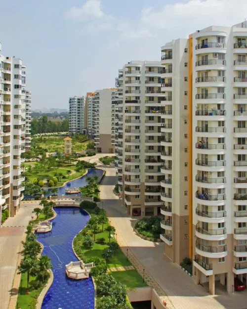 Aerial view of BrandLand's BTL marketing activation event in a lush residential apartment complex in India, featuring tall modern buildings, a winding blue canal, green landscapes, and on-ground promotional setup by BrandLand.